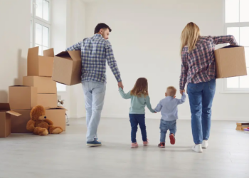 Two adults and two young children walk into a room holding hands, surrounded by moving boxes and a teddy bear, suggesting they are moving into a new home.