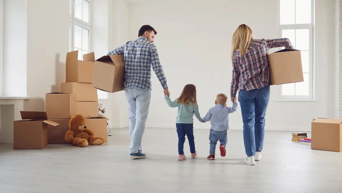 Two adults and two young children walk into a room holding hands, surrounded by moving boxes and a teddy bear, suggesting they are moving into a new home.