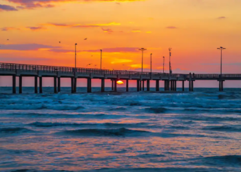 Sunset glows orange behind a long seaside pier, with silhouetted lamps and birds above rolling blue waves; a serene coastal scene.