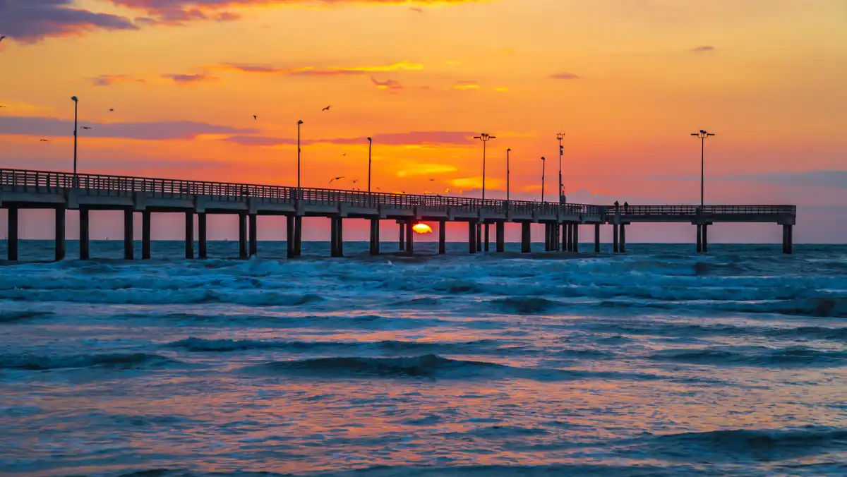 Sunset glows orange behind a long seaside pier, with silhouetted lamps and birds above rolling blue waves; a serene coastal scene.