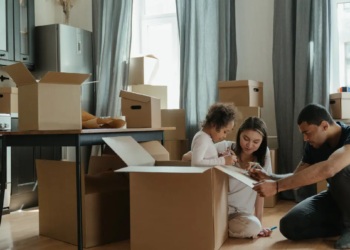 Family unpacking cardboard moving boxes in a bright living room; parents help their young daughter, creating a warm, joyful moving day.