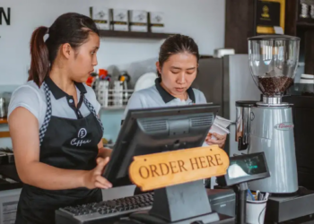 Two baristas work at a café counter beside a coffee grinder and register marked “ORDER HERE,” conveying focused, welcoming service.