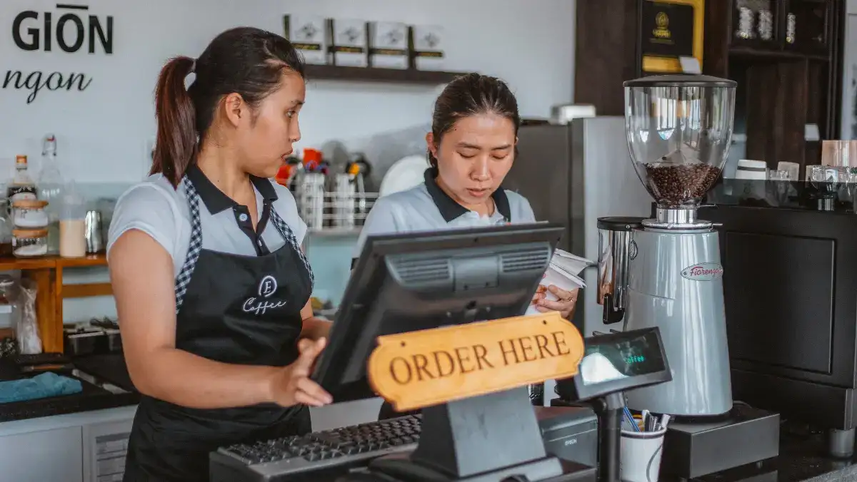 Two baristas work at a café counter beside a coffee grinder and register marked “ORDER HERE,” conveying focused, welcoming service.