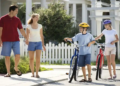 Parents walk with two helmeted children and their bicycles on a sunny suburban sidewalk, creating a cheerful family scene by a white picket fence.
