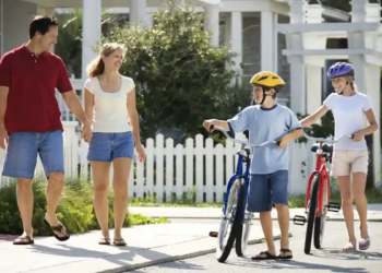 Parents walk with two helmeted children and their bicycles on a sunny suburban sidewalk, creating a cheerful family scene by a white picket fence.