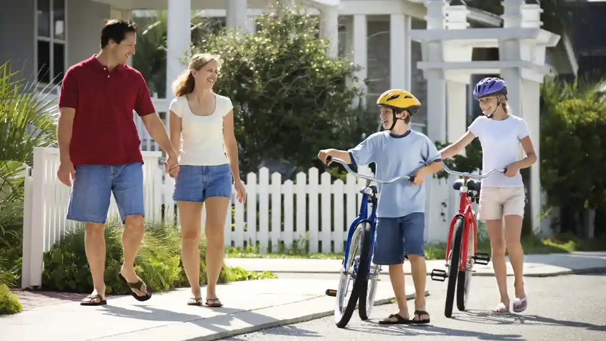 Parents walk with two helmeted children and their bicycles on a sunny suburban sidewalk, creating a cheerful family scene by a white picket fence.