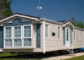 Beige mobile home with blue-gray trim, large bay windows, white steps, and a blue canopy, set on a sunny grassy lot beneath a clear sky.