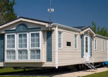 Beige mobile home with blue-gray trim, large bay windows, white steps, and a blue canopy, set on a sunny grassy lot beneath a clear sky.