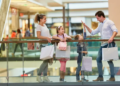 Family shopping at a mall: parents and two children stand by a glass railing, holding bags and smiling in a bright, cheerful setting.