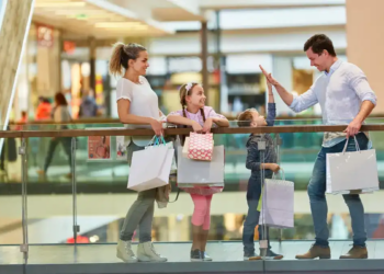 Family shopping at a mall: parents and two children stand by a glass railing, holding bags and smiling in a bright, cheerful setting.