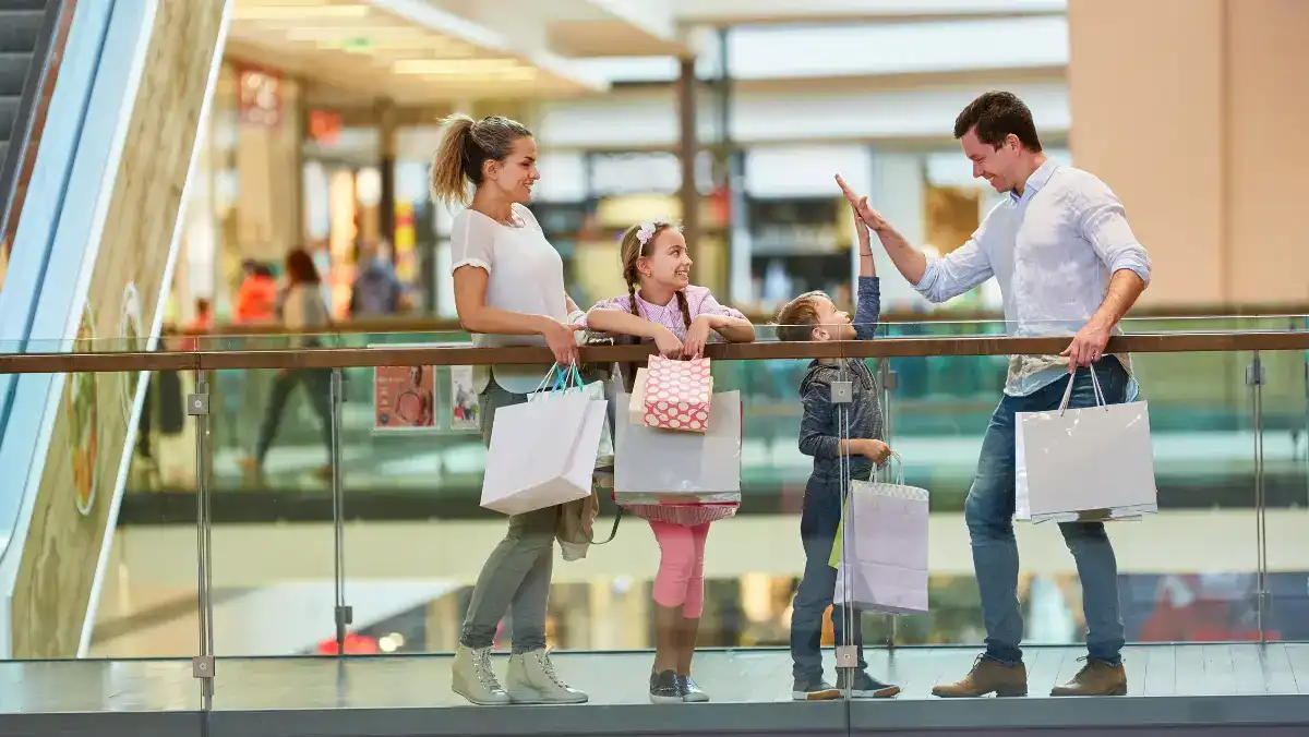 Family shopping at a mall: parents and two children stand by a glass railing, holding bags and smiling in a bright, cheerful setting.
