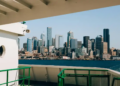 View from a ferry deck toward Seattle’s modern skyline across Elliott Bay, framed by green railings under a bright, clear blue sky.