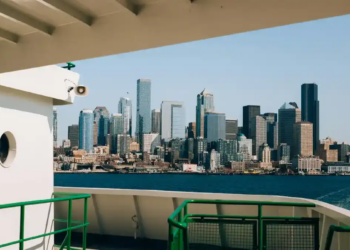 View from a ferry deck toward Seattle’s modern skyline across Elliott Bay, framed by green railings under a bright, clear blue sky.